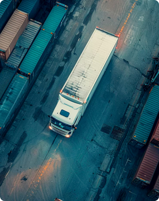 Aerial view of a white semi-truck driving through a shipping container yard with rows of colorful containers on each side.